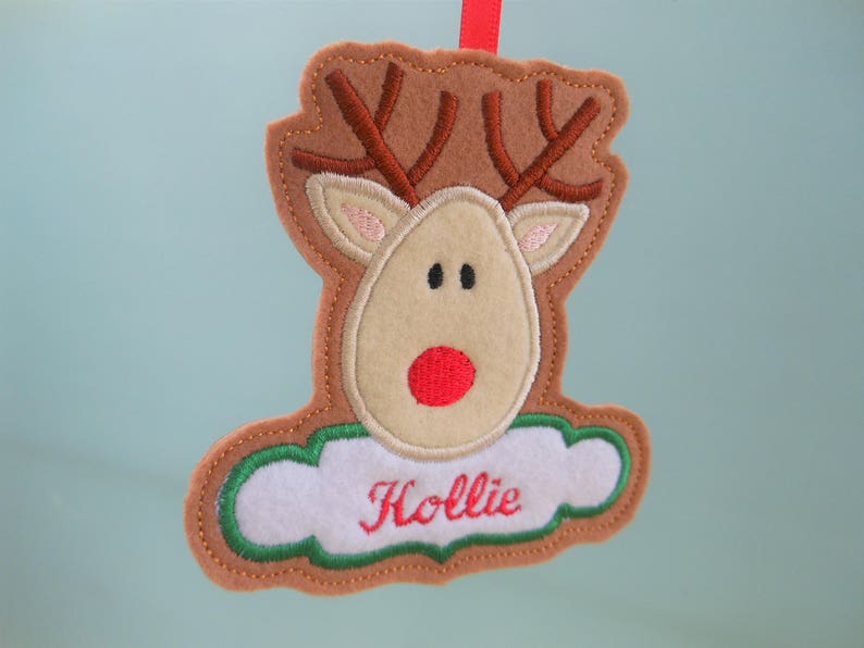 K&ouml;nnte beinhalten: Ein Weihnachtsornament aus Filz in Form eines Rentiers. Es hat braune H&ouml;rner, ein beiges Gesicht und eine rote Nase. Der Name "Hollie" ist rot auf einem wei&szlig;en, wolkenf&ouml;rmigen Bereich gestickt.