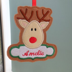 K&ouml;nnte beinhalten: Ein Weihnachtsornament aus Filz in Form eines Rentiers mit braunem Geweih, beigem Gesicht und roter Nase. Der Name "Amelie" ist rot auf einer wei&szlig;en, wolkenf&ouml;rmigen Basis mit gr&uuml;nen R&auml;ndern gestickt. Ein rotes Band zum Aufh&auml;ngen.