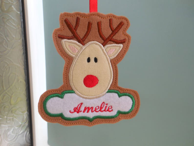 K&ouml;nnte beinhalten: Ein Weihnachtsornament aus Filz in Form eines Rentiers mit braunem Geweih, beigem Gesicht und roter Nase. Der Name "Amelie" ist rot auf einer wei&szlig;en, wolkenf&ouml;rmigen Basis mit gr&uuml;nen R&auml;ndern gestickt. Ein rotes Band zum Aufh&auml;ngen.