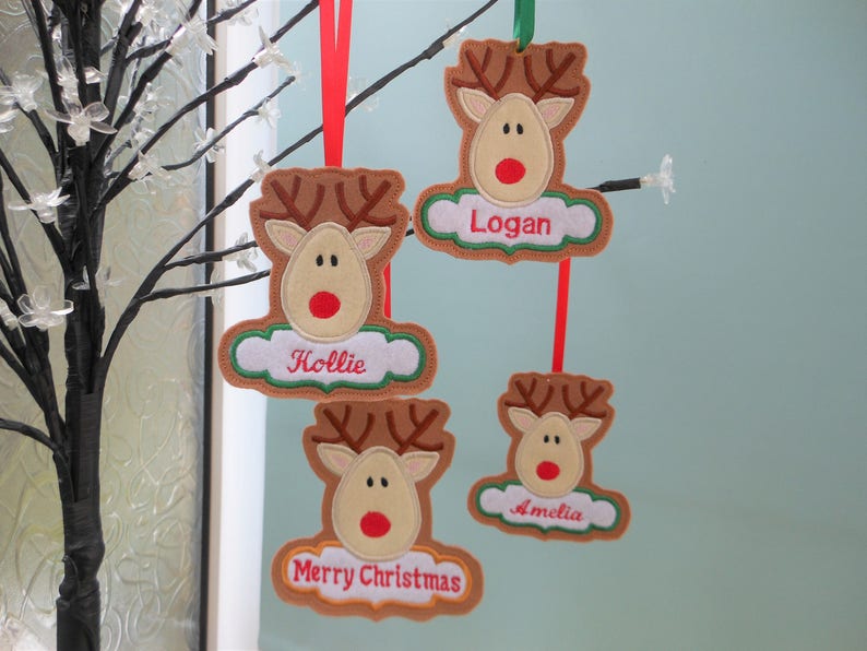 K&ouml;nnte beinhalten: Weihnachtsschmuck in Form von Rentieren mit Namen und "Frohe Weihnachten". Die Rentiere sind braun mit beigen Gesichtern, roten Nasen und gr&uuml;n-wei&szlig;en Wolken-Namensschildern. Sie h&auml;ngen an roten und gr&uuml;nen B&auml;ndern.