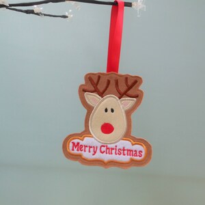 K&ouml;nnte beinhalten: Ein Weihnachtsornament aus Filz in Form eines Rentiers mit braunem Geweih, beigem Gesicht und roter Nase. Das Ornament tr&auml;gt die gestickten Worte "Merry Christmas" und wird von einem roten Band gehalten.
