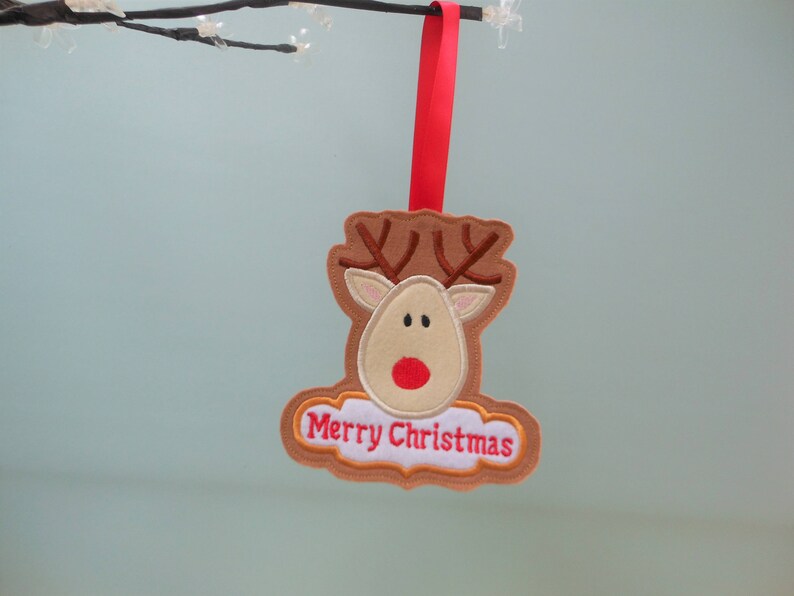 K&ouml;nnte beinhalten: Ein Weihnachtsornament aus Filz in Form eines Rentiers mit braunem Geweih, beigem Gesicht und roter Nase. Das Ornament tr&auml;gt die gestickten Worte "Merry Christmas" und wird von einem roten Band gehalten.