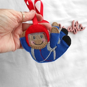 May include: A felt skydiver ornament with a blue jumpsuit and red helmet. The skydiver has a smiling face and silver detailing. It has a red ribbon for hanging and is designed for crafts.