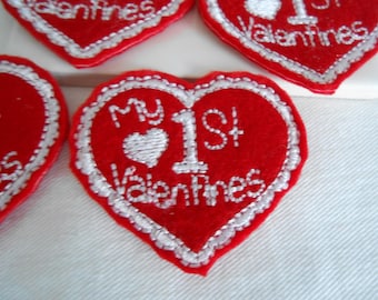 Może przedstawiać: Czerwone naszywki w kształcie serca z białym haftowanym wykończeniem i tekstem. Na naszywkach widnieje napis "My 1st Valentines" i małe białe serce. Te naszywki są prawdopodobnie na Walentynki.