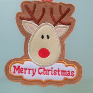 K&ouml;nnte beinhalten: Ein Weihnachtsornament aus Filz in Form eines Rentierkopfes. Das Rentier hat braune Geweihe, ein beiges Gesicht und eine rote Nase. Die Worte "Merry Christmas" sind rot auf einem wei&szlig;en, wolkenf&ouml;rmigen Banner gestickt.