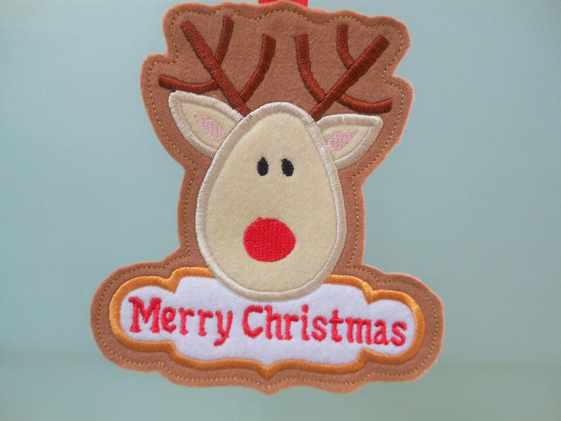 K&ouml;nnte beinhalten: Ein Weihnachtsornament aus Filz in Form eines Rentierkopfes. Das Rentier hat braune Geweihe, ein beiges Gesicht und eine rote Nase. Die Worte "Merry Christmas" sind rot auf einem wei&szlig;en, wolkenf&ouml;rmigen Banner gestickt.