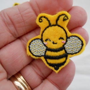May include: A yellow felt bee applique with black stripes and embroidered wings. The bee has a smiling face with pink cheeks. The applique is held in a hand.