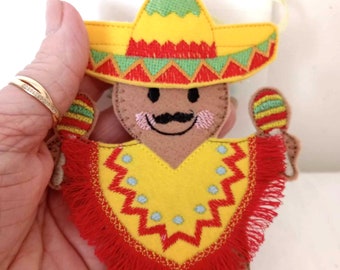 Może przedstawiać: Filcowa ozdoba w kształcie piernikowego ludzika w żółtym sombrero i ponczo. Sombrero ma zielone i czerwone detale. Ponczo jest żółte z czerwonymi frędzlami i niebieskimi akcentami. Piernikowy ludzik ma czarne wąsy i trzyma marakasy.