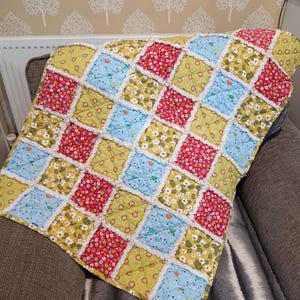 May include: A patchwork quilt featuring squares of floral fabric in red, yellow, and blue. The quilt has a fringed edge and is draped over a grey armchair. The design includes small white flowers and other decorative elements, creating a cozy, handmade feel.