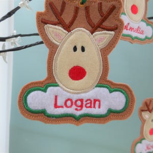 K&ouml;nnte beinhalten: Filz-Rentier-Ornament mit dem Namen "Logan" in roter Schrift auf einem wei&szlig;en, wolkenf&ouml;rmigen Anh&auml;nger. Das Rentier hat braunes Geweih, ein beiges Gesicht und eine rote Nase. Weitere Ornamente sind sichtbar.