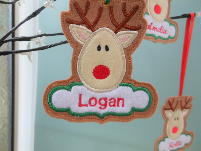 K&ouml;nnte beinhalten: Filz-Rentier-Ornament mit dem Namen "Logan" in roter Schrift auf einem wei&szlig;en, wolkenf&ouml;rmigen Anh&auml;nger. Das Rentier hat braunes Geweih, ein beiges Gesicht und eine rote Nase. Weitere Ornamente sind sichtbar.