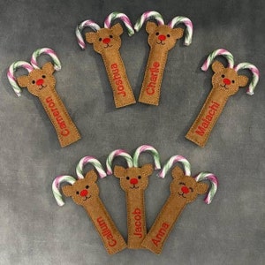 May include: A selection of reindeer-shaped candy cane holders. Each holder is crafted from brown felt and features a reindeer face with a red nose. The holders are personalised with names in red embroidery, and paired with candy canes.