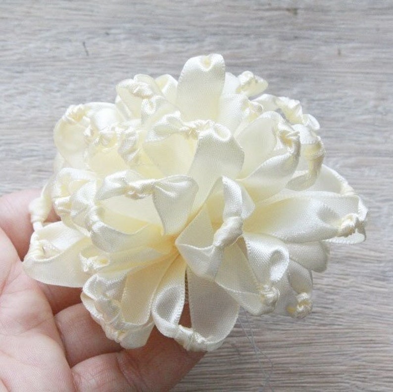 K&ouml;nnte beinhalten: Eine cremefarbene Satinbandblume mit einer gekr&auml;uselten Textur. Die Blume wird durch Drehen und Falten des Bandes hergestellt, um eine dreidimensionale Form zu erzeugen.