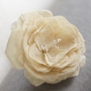 Könnte beinhalten: Eine zarte, cremefarbene Stoffblume mit einem zentralen Detail aus kleinen, weißen Perlen.