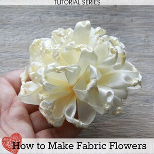 K&ouml;nnte beinhalten: Nahaufnahme einer wei&szlig;en Stoffblume, die durch das Knoten von Band hergestellt wurde. Die Blume hei&szlig;t "Geknotete Chrysantheme" und ist Teil einer Tutorial-Serie namens "Jewel Box Ballerina".