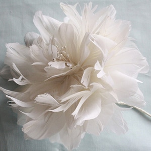 Peut inclure: Une fleur blanche en plumes avec un embellissement de perles et de strass. La fleur est faite de plumes et a un aspect délicat et aérien.