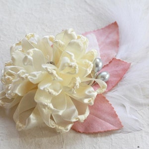 K&ouml;nnte beinhalten: Eine zarte, elfenbeinfarbene Satinblume mit gekr&auml;uselter Textur, akzentuiert mit zwei perlmuttartigen Perlen und rosa Stoffbl&auml;ttern. Die Blume ist von wei&szlig;en Federn umgeben.