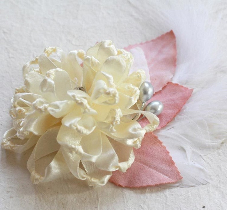 K&ouml;nnte beinhalten: Eine zarte, elfenbeinfarbene Satinblume mit gekr&auml;uselter Textur, akzentuiert mit zwei perlmuttartigen Perlen und rosa Stoffbl&auml;ttern. Die Blume ist von wei&szlig;en Federn umgeben.