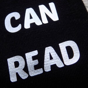 Op de afbeelding: Een zwarte stoffen achtergrond met de woorden "CAN READ" gedrukt in witte reflecterende letters.