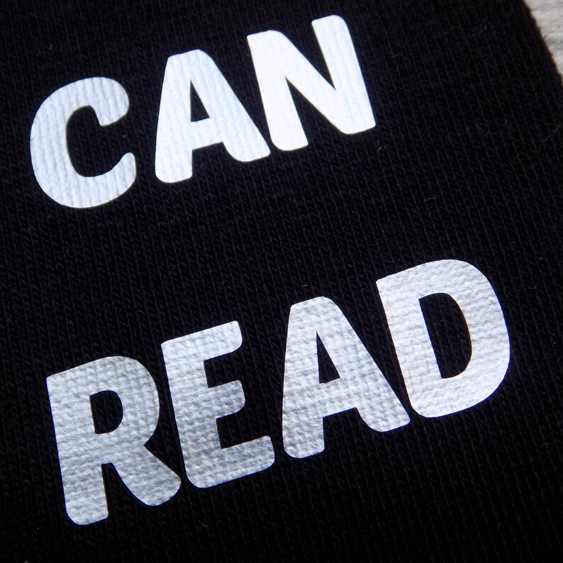 Op de afbeelding: Een zwarte stoffen achtergrond met de woorden "CAN READ" gedrukt in witte reflecterende letters.