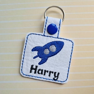 May include: White leather keyring with a blue rocket ship and the name "Harry" embroidered on it. The keyring has a blue button and a silver key ring.