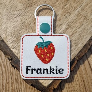 May include: White leather keychain with a red embroidered strawberry and the name Frankie in black embroidery.
