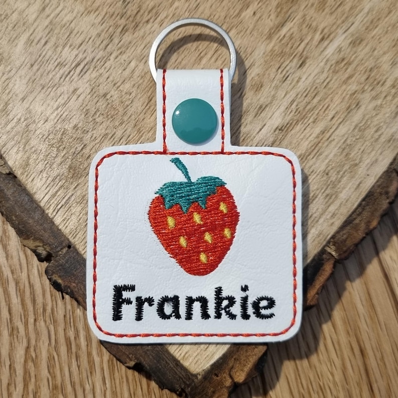 May include: White leather keychain with a red embroidered strawberry and the name Frankie in black embroidery.