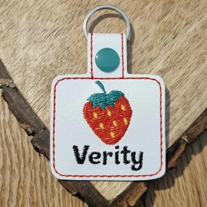 May include: White leather keychain with a red embroidered strawberry and the name "Verity" in black embroidery.