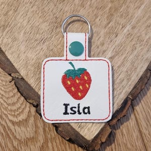 May include: White leather keychain with a red and yellow embroidered strawberry and the name Isla in black embroidery.