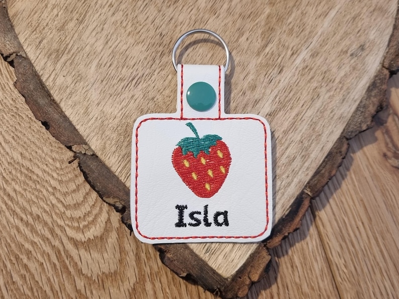 May include: White leather keychain with a red and yellow embroidered strawberry and the name Isla in black embroidery.