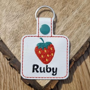 May include: White leather keychain with a red embroidered strawberry and the name "Ruby" in black embroidery.
