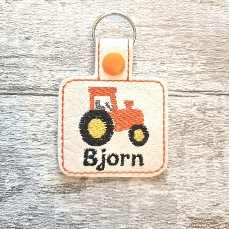 May include: A white leather keychain with an orange tractor design and the name "Bjorn" embroidered in black thread.