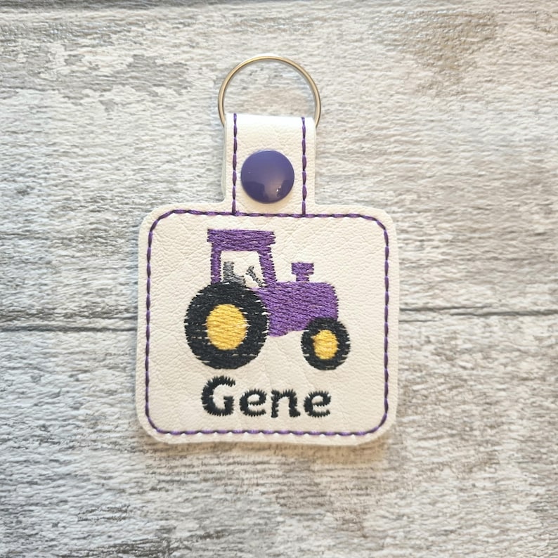 May include: Purple tractor keychain with a white background and the name "Gene" embroidered on the bottom.
