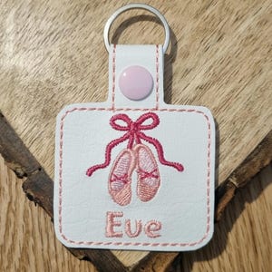 May include: White leather keychain with a pink embroidered ballerina shoe design and the name "Eve" stitched below.