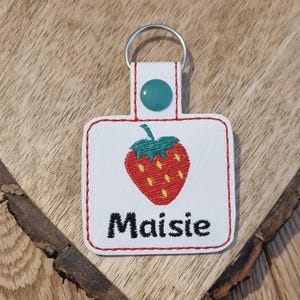 May include: White fabric keychain with a red embroidered strawberry and the name "Maisie" in black embroidery.
