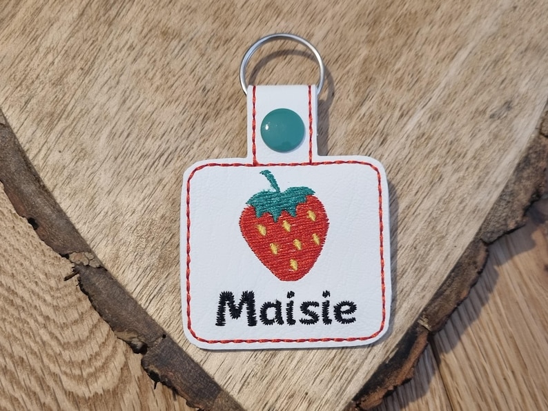May include: White fabric keychain with a red embroidered strawberry and the name "Maisie" in black embroidery.