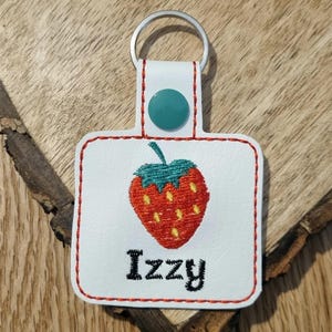 May include: White leather keychain with a red embroidered strawberry and the name "Izzy" in black embroidery. The keychain has a teal button and a silver key ring.