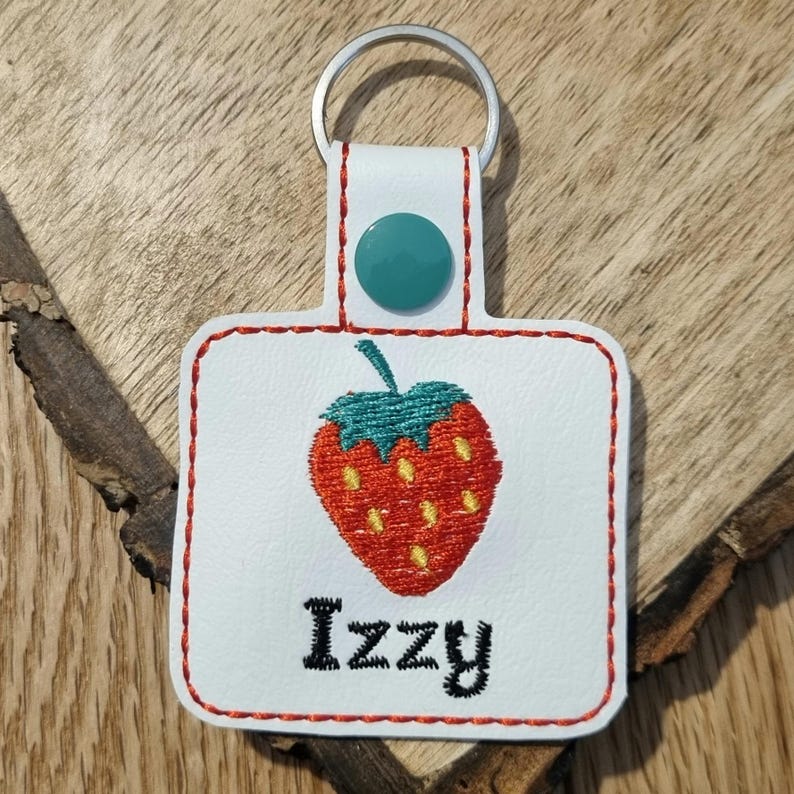 May include: White leather keychain with a red embroidered strawberry and the name "Izzy" in black embroidery. The keychain has a teal button and a silver key ring.