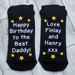 Puede incluir: Un par de calcetines negros con texto blanco y estrellas amarillas. Un calcet&iacute;n dice "Happy Birthday to the Best Daddy!" y el otro calcet&iacute;n dice "Love Finlay and Henry xxx".