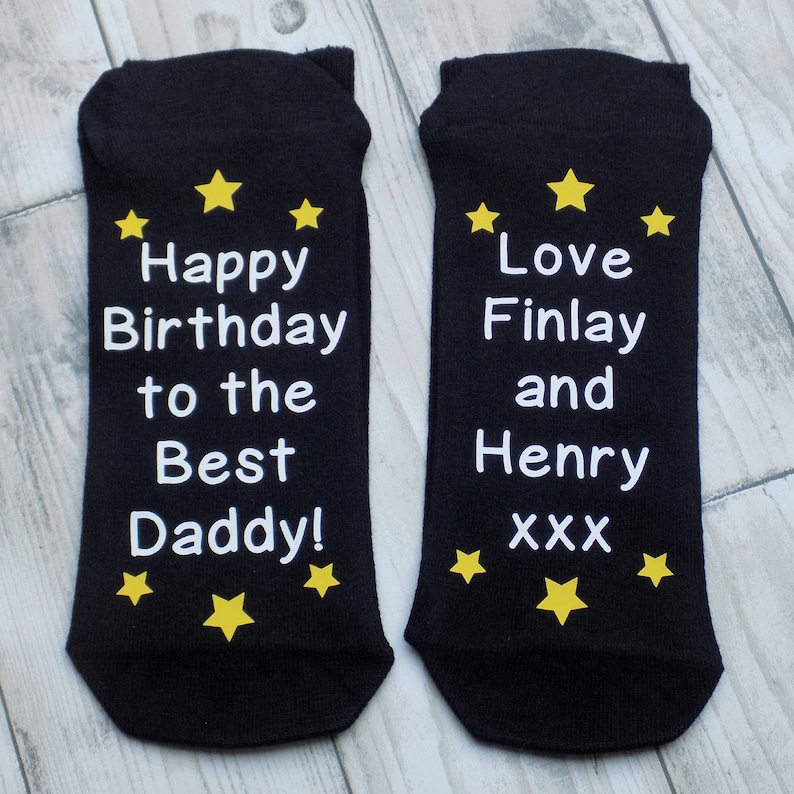 Puede incluir: Un par de calcetines negros con texto blanco y estrellas amarillas. Un calcet&iacute;n dice "Happy Birthday to the Best Daddy!" y el otro calcet&iacute;n dice "Love Finlay and Henry xxx".