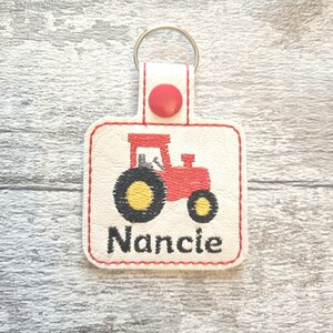 May include: White leather keychain with a red tractor design and the name "Nancie" embroidered in black thread.