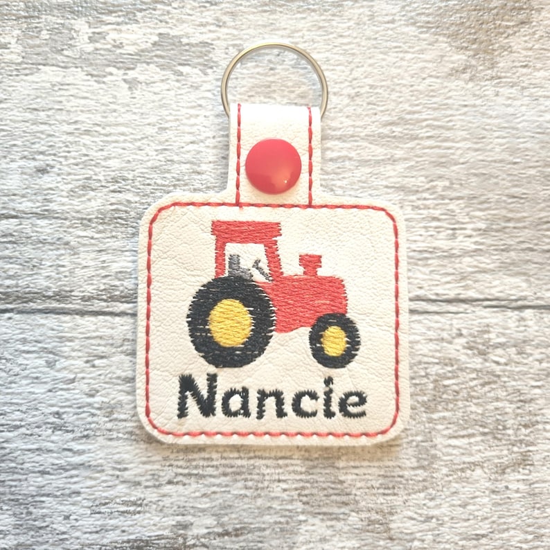 May include: White leather keychain with a red tractor design and the name "Nancie" embroidered in black thread.