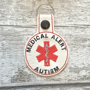 May include: White leather keychain with a red medical symbol and the text "MEDICAL ALERT AUTISM".