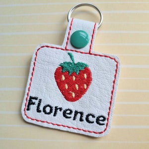 May include: White leather keychain with a red embroidered strawberry and the name Florence in black embroidery. The keychain has a silver key ring and a teal button.