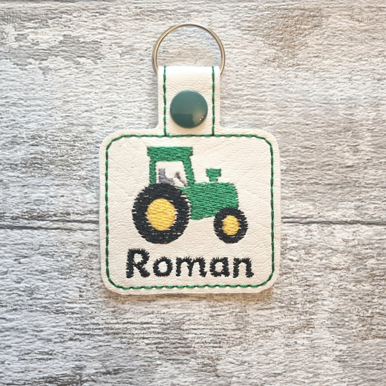 May include: A white leather keychain with a green tractor design and the name "Roman" embroidered in green thread.