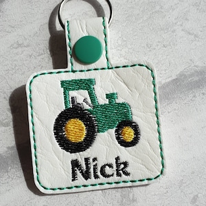 May include: Green and black embroidered tractor keychain with the name Nick on a white leather background.