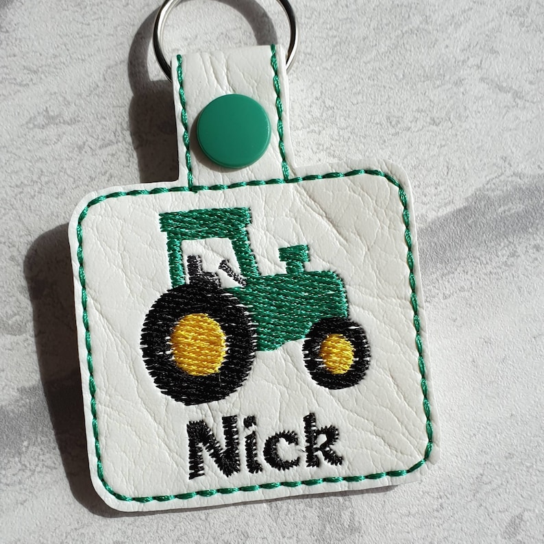 May include: Green and black embroidered tractor keychain with the name Nick on a white leather background.