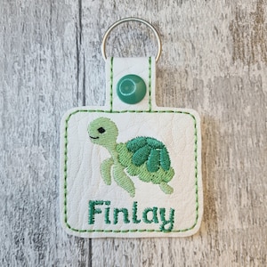 May include: White square keychain featuring a green embroidered turtle and the name "Finlay" in green. It has a green button and a silver key ring. The edges are stitched in green.
