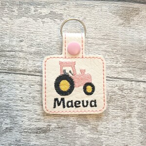 May include: Pink tractor keychain with a white background and the name "Maeva" embroidered in black.