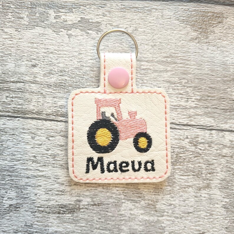May include: Pink tractor keychain with a white background and the name "Maeva" embroidered in black.